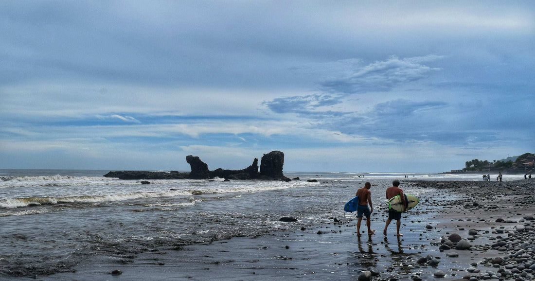 Two surfers walking on a black sand beach with rock formations in El Salvador