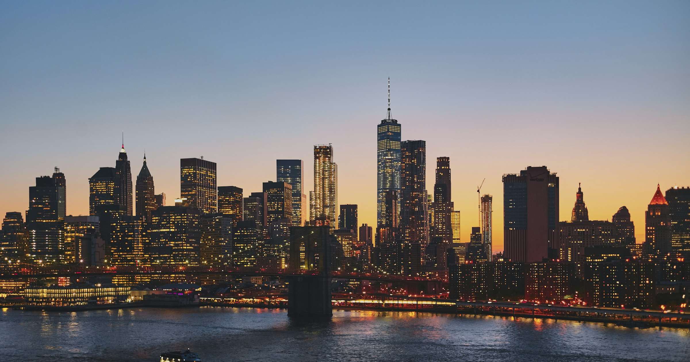 New York City skyline at dusk, with Brooklyn Bridge and Manhattan towers reflected in the East River
